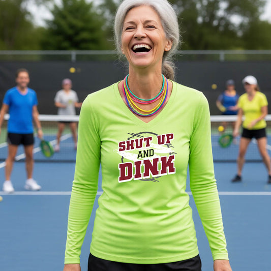 A woman on a pickleball court wearing a lime green long-sleeve t-shirt with a white, gray and black graphic of the text: ‘Shut Up and Dink, standing on a pickleball court.