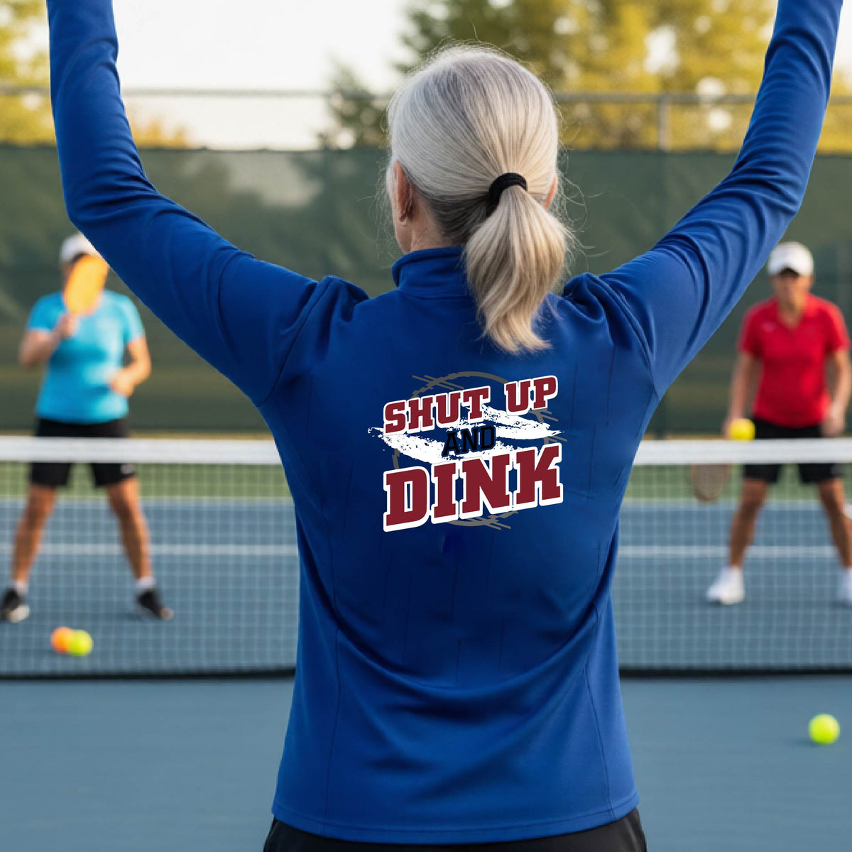 A woman on a pickleball court, facing backwards, wearing a royal blue quarter-zip shirt with a white, gray and black graphic of the text: ‘Shut Up and Dink, standing on a pickleball court.