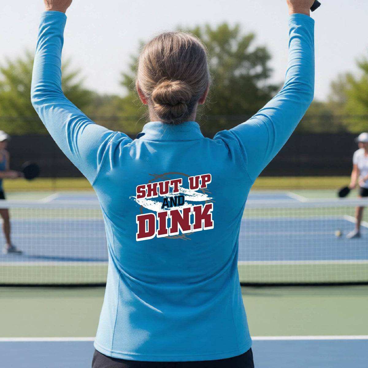 A woman on a pickleball court, facing backwards, wearing a bright blue quarter-zip shirt with a white, gray and black graphic of the text: ‘Shut Up and Dink, standing on a pickleball court. 
