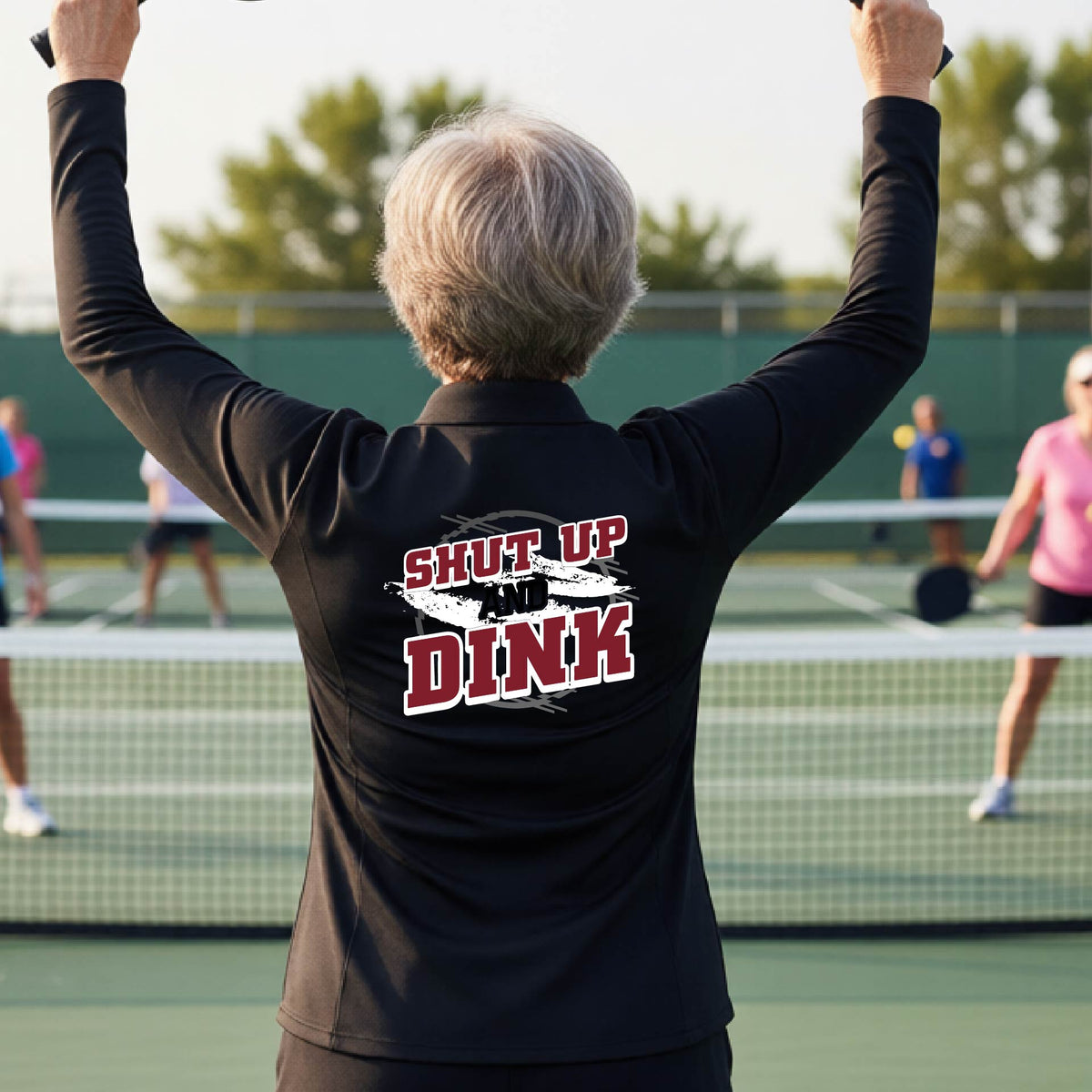 A woman on a pickleball court, facing backwards, wearing a black quarter-zip shirt with a white, gray and black graphic of the text: ‘Shut Up and Dink, standing on a pickleball court.