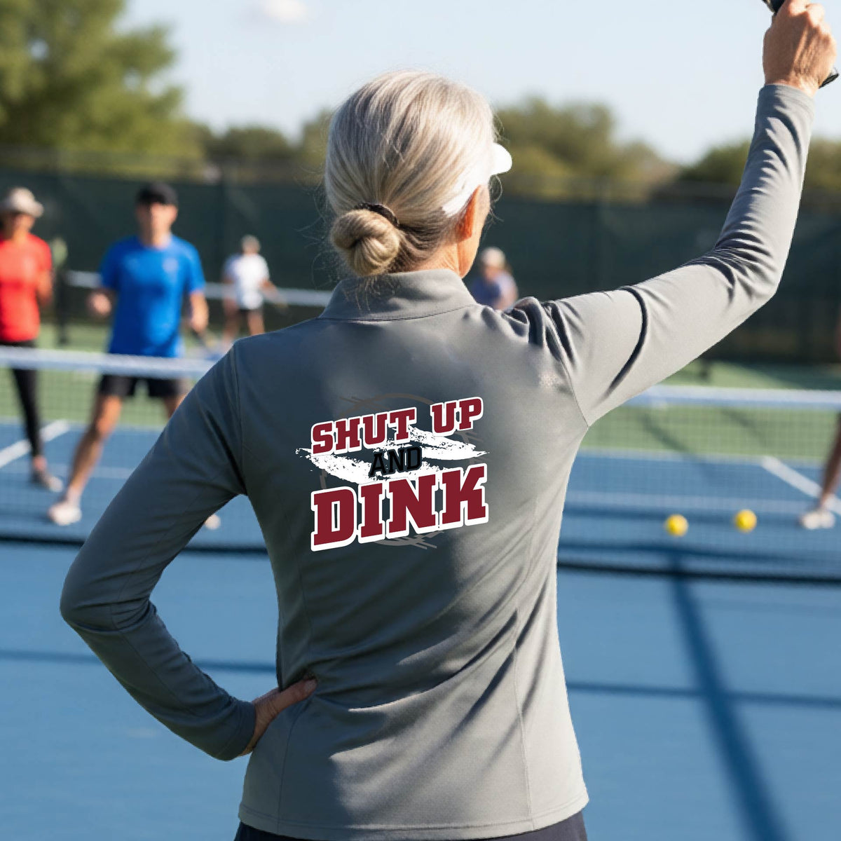 A woman on a pickleball court, facing backwards, wearing a dark gray quarter-zip shirt with a white, gray and black graphic of the text: ‘Shut Up and Dink, standing on a pickleball court.