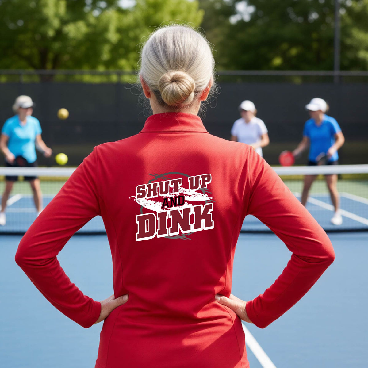 A woman on a pickleball court, facing backwards, wearing a red quarter-zip shirt with a white, gray and black graphic of the text: ‘Shut Up and Dink, standing on a pickleball court.