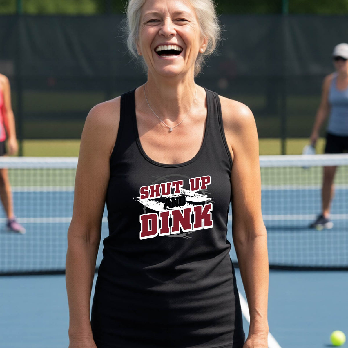 A woman on a pickleball court wearing a black racerback tank topwith a white, gray and black graphic of the text: ‘Shut Up and Dink, standing on a pickleball court.