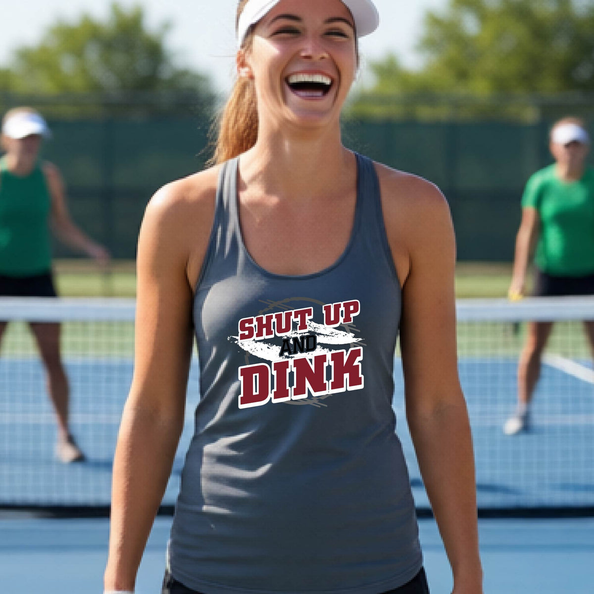 A woman on a pickleball court wearing a dark gray racerback tank topwith a white, gray and black graphic of the text: ‘Shut Up and Dink, standing on a pickleball court.
