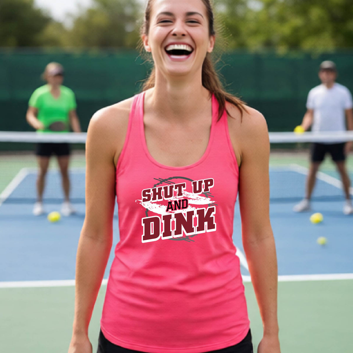 A woman on a pickleball court wearing a bright coral pink racerback tank topwith a white, gray and black graphic of the text: ‘Shut Up and Dink, standing on a pickleball court.