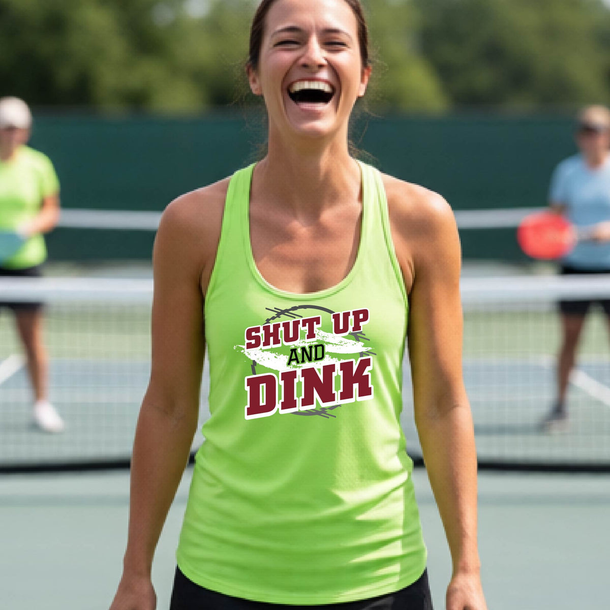 A woman on a pickleball court wearing a lime green racerback tank topwith a white, gray and black graphic of the text: ‘Shut Up and Dink, standing on a pickleball court.