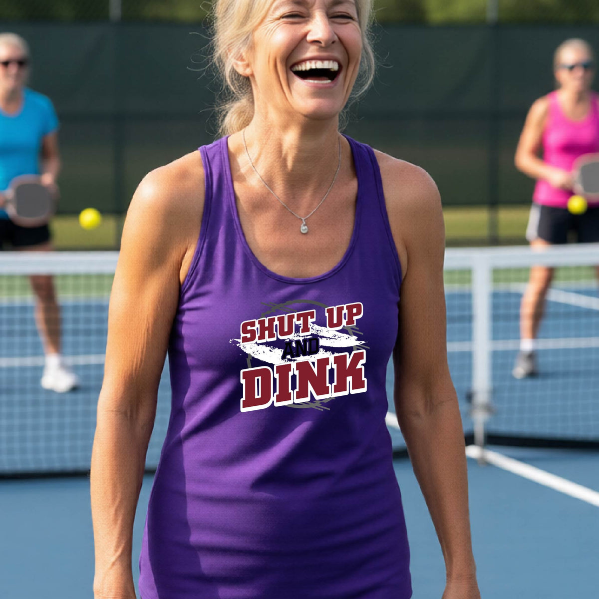 A woman on a pickleball court wearing a purple racerback tank topwith a white, gray and black graphic of the text: ‘Shut Up and Dink, standing on a pickleball court.
