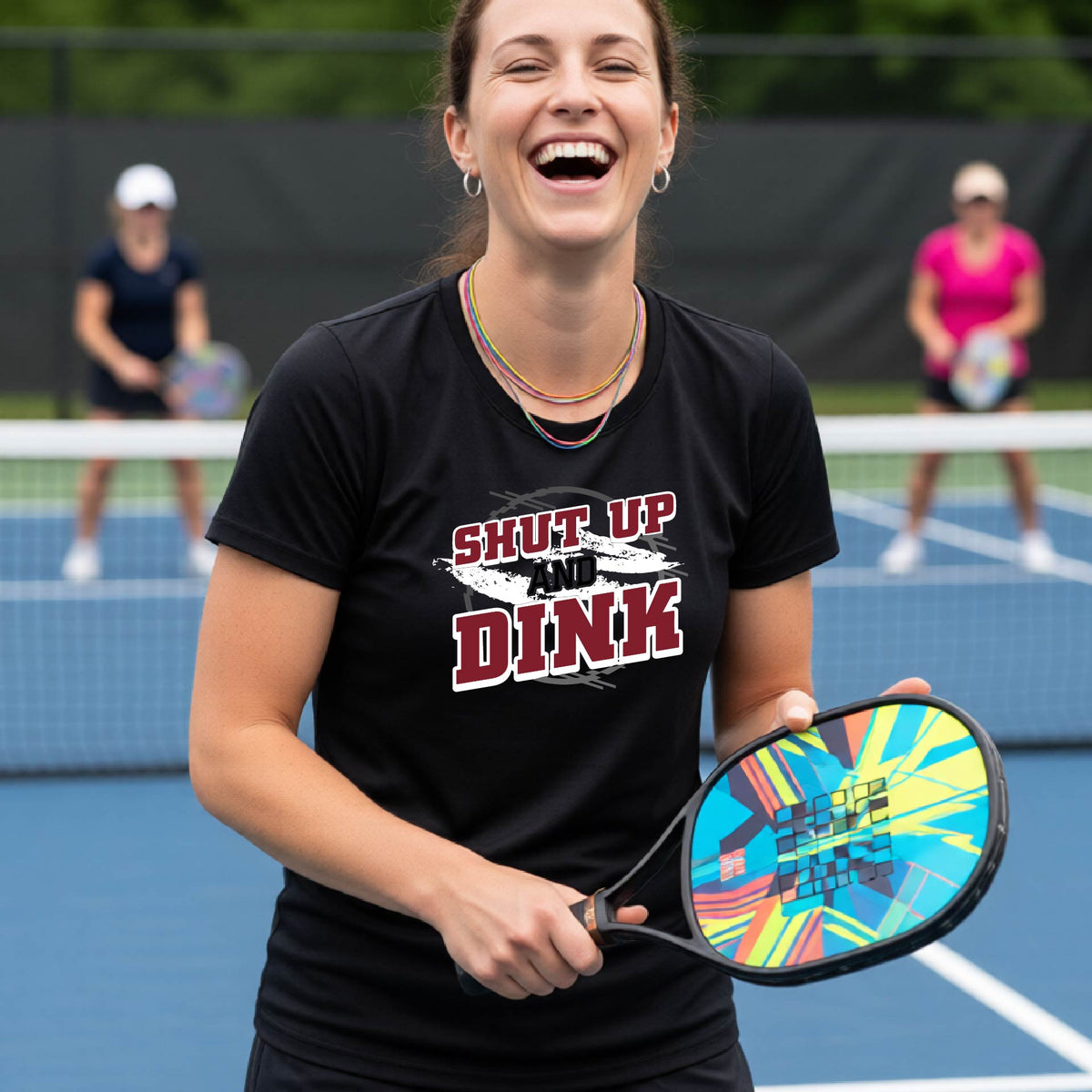 A woman on a pickleball court wearing a black short-sleeve t-shirt with a white, gray and black graphic of the text: ‘Shut Up and Dink, standing on a pickleball court.