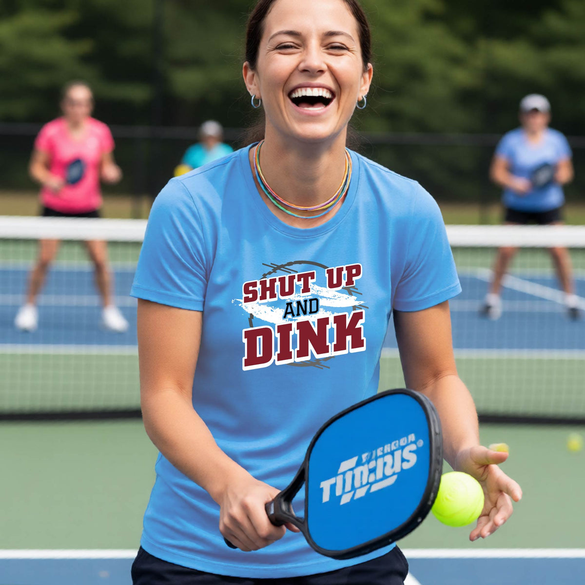 A woman on a pickleball court wearing a blue short-sleeve t-shirt with a white, gray and black graphic of the text: ‘Shut Up and Dink, standing on a pickleball court.