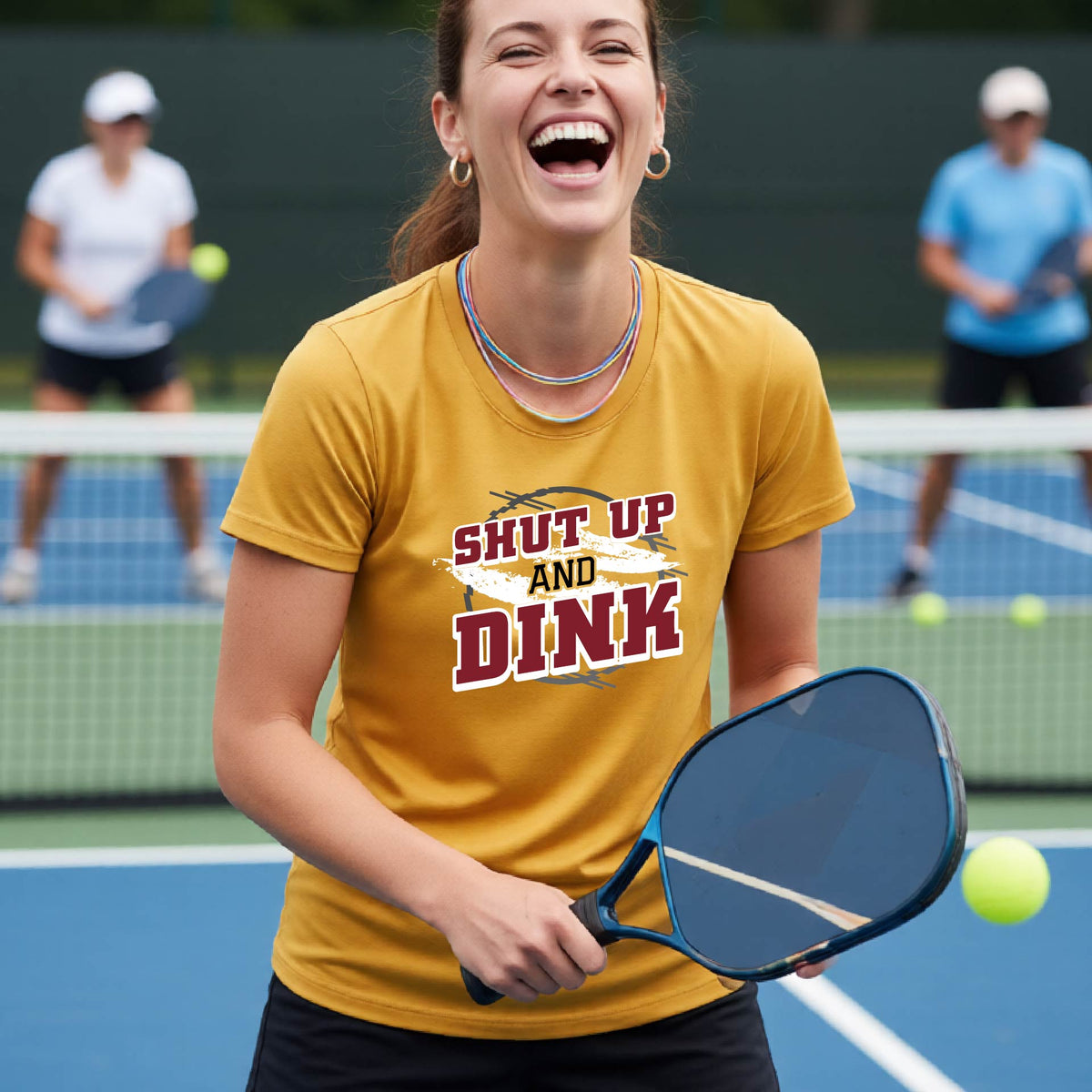 A woman on a pickleball court wearing a mustard yellow gold short-sleeve t-shirt with a white, gray and black graphic of the text: ‘Shut Up and Dink, standing on a pickleball court.