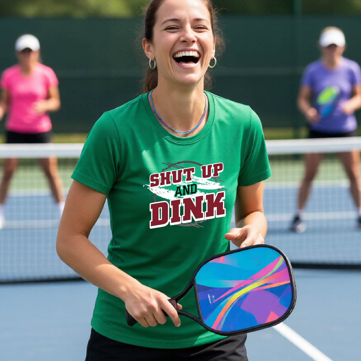 A woman on a pickleball court wearing a green short-sleeve t-shirt with a white, gray and black graphic of the text: ‘Shut Up and Dink, standing on a pickleball court.