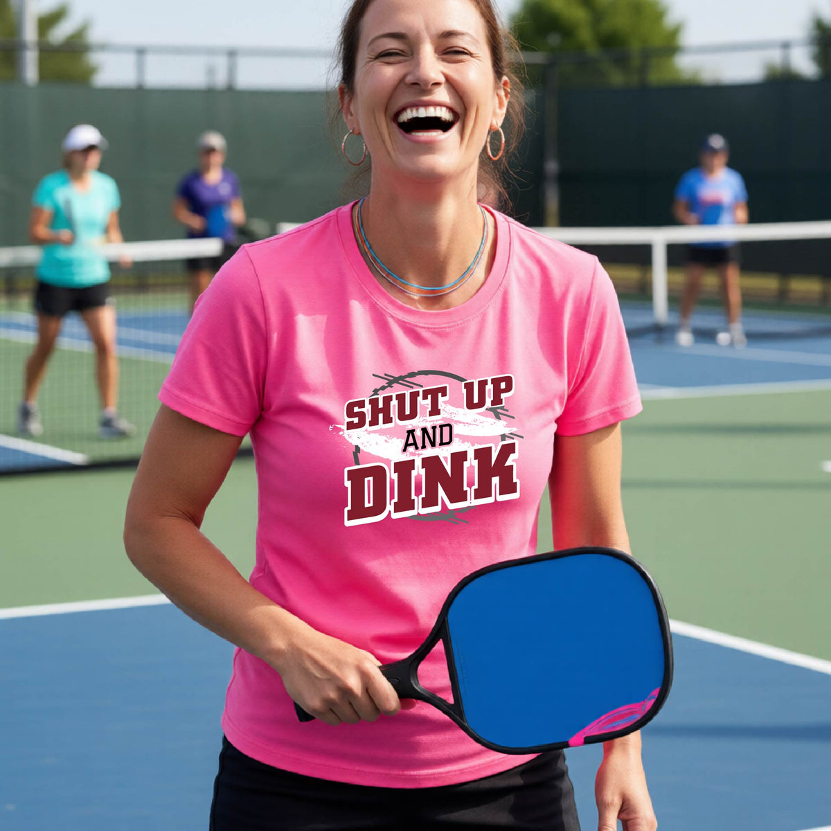A woman on a pickleball court wearing a pink short-sleeve t-shirt with a white, gray and black graphic of the text: ‘Shut Up and Dink, standing on a pickleball court.