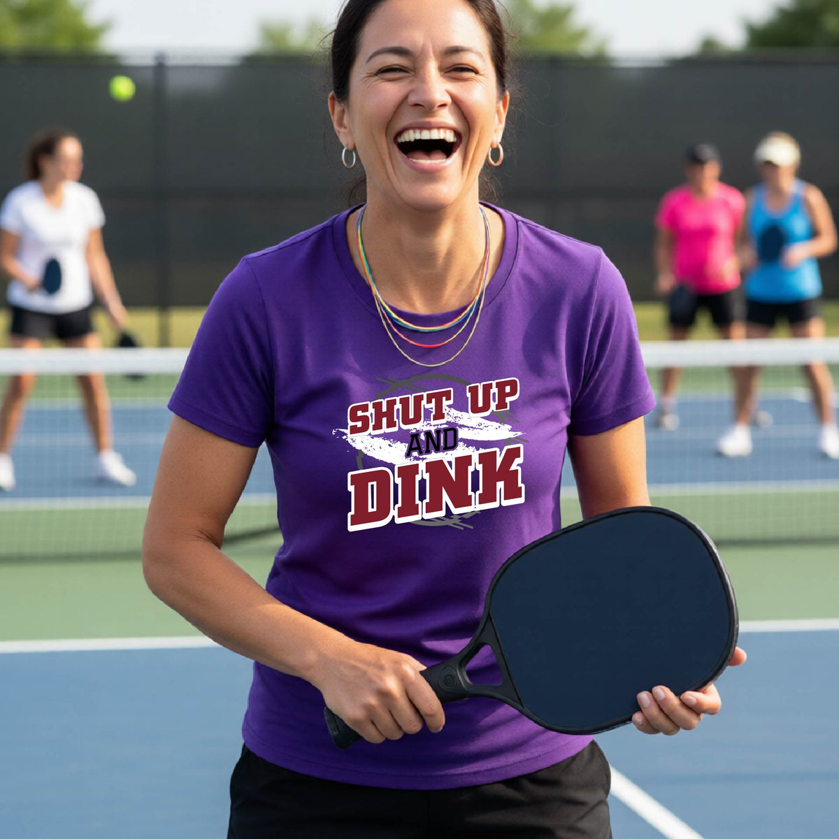 A woman on a pickleball court wearing a purple short-sleeve t-shirt with a white, gray and black graphic of the text: ‘Shut Up and Dink, standing on a pickleball court.