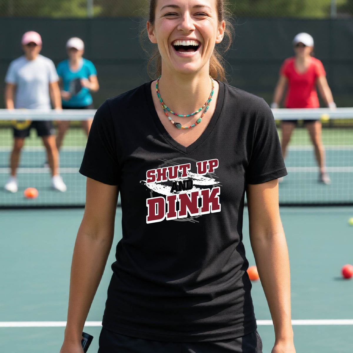 A woman on a pickleball court wearing a black short-sleeve v-neck t-shirt with a white, gray and black graphic of the text: ‘Shut Up and Dink, standing on a pickleball court.