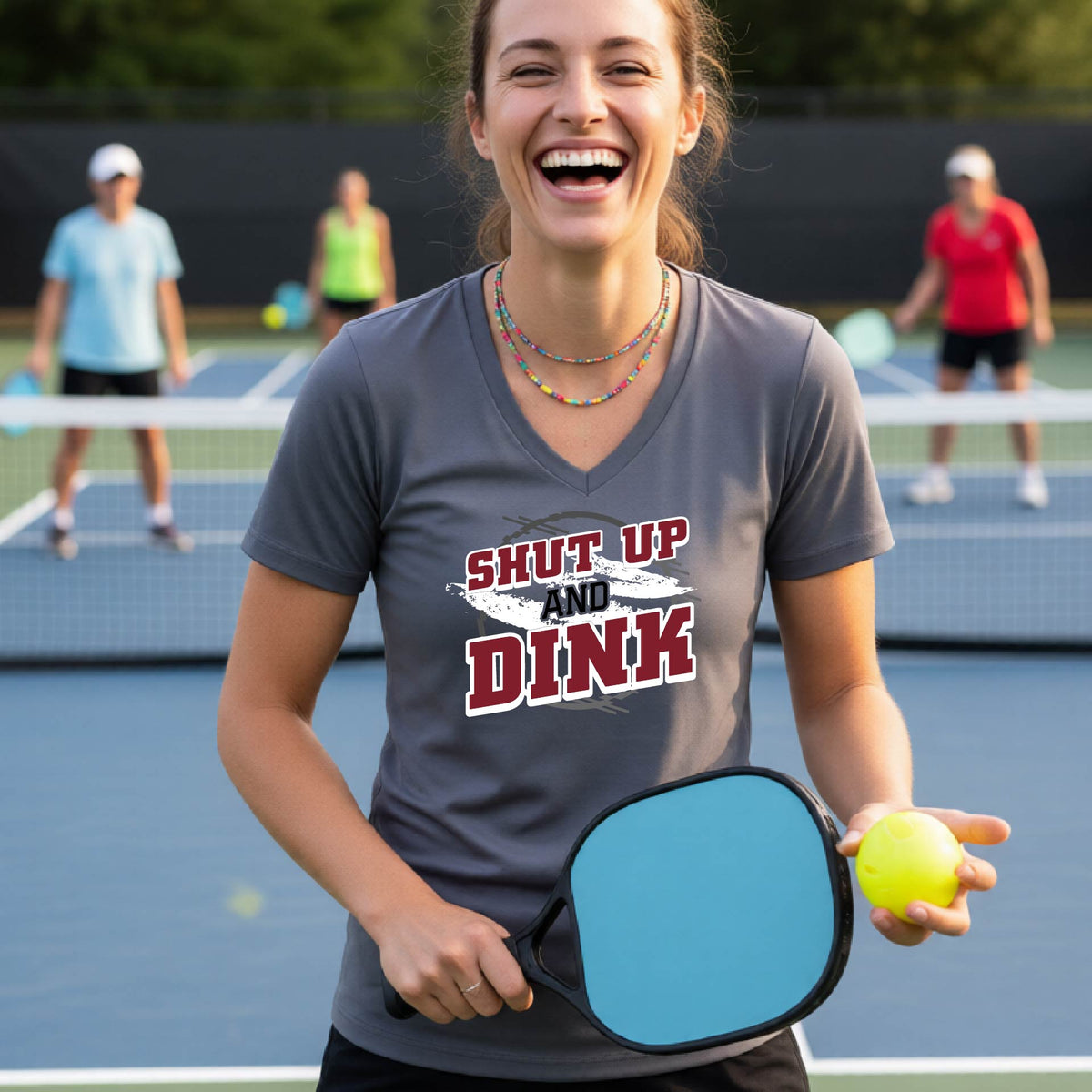 A woman on a pickleball court wearing a dark gray short-sleeve v-neck t-shirt with a white, gray and black graphic of the text: ‘Shut Up and Dink, standing on a pickleball court.