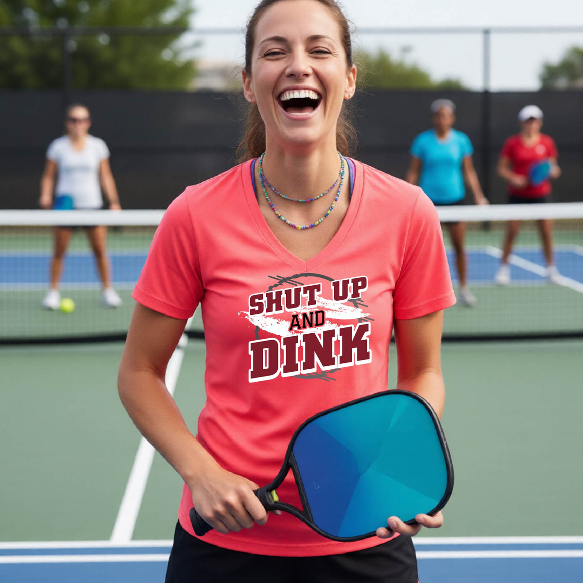 A woman on a pickleball court wearing a bright coral pink short-sleeve v-neck t-shirt with a white, gray and black graphic of the text: ‘Shut Up and Dink, standing on a pickleball court.