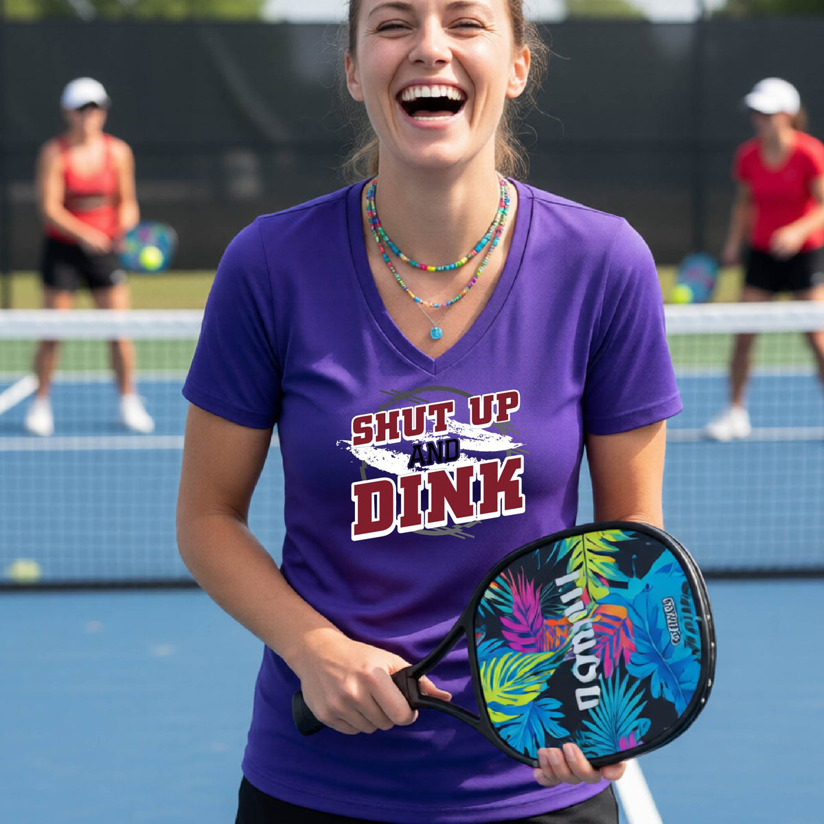 A woman on a pickleball court wearing a purple short-sleeve v-neck t-shirt with a white, gray and black graphic of the text: ‘Shut Up and Dink, standing on a pickleball court.
