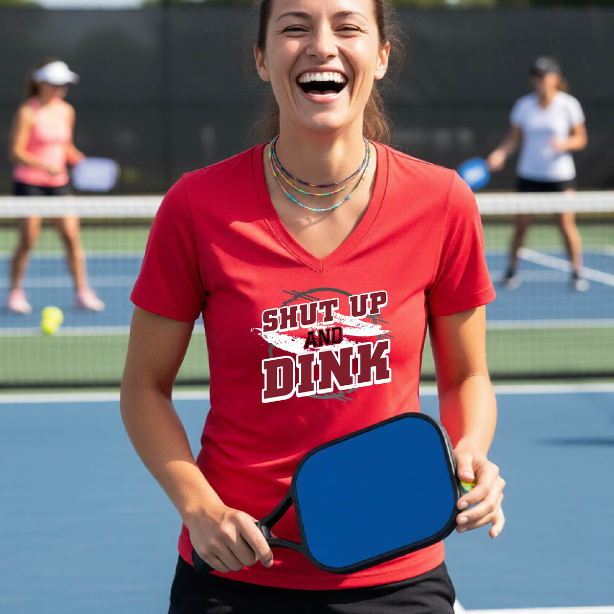 A woman on a pickleball court wearing a red short-sleeve v-neck t-shirt with a white, gray and black graphic of the text: ‘Shut Up and Dink, standing on a pickleball court.