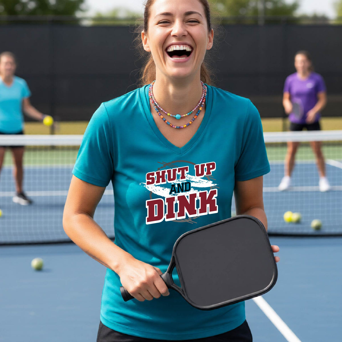 A woman on a pickleball court wearing a teal short-sleeve v-neck t-shirt with a white, gray and black graphic of the text: ‘Shut Up and Dink, standing on a pickleball court.