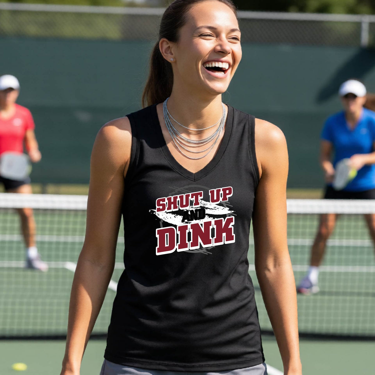 A woman on a pickleball court wearing a black sleeveless t-shirt with a white, gray and black graphic of the text: ‘Shut Up and Dink, standing on a pickleball court.