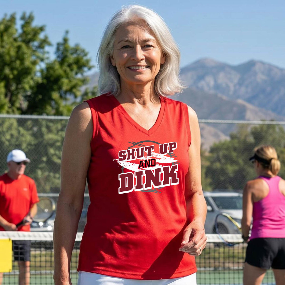 A woman on a pickleball court wearing a red sleeveless t-shirt with a white, gray and black graphic of the text: ‘Shut Up and Dink, standing on a pickleball court.