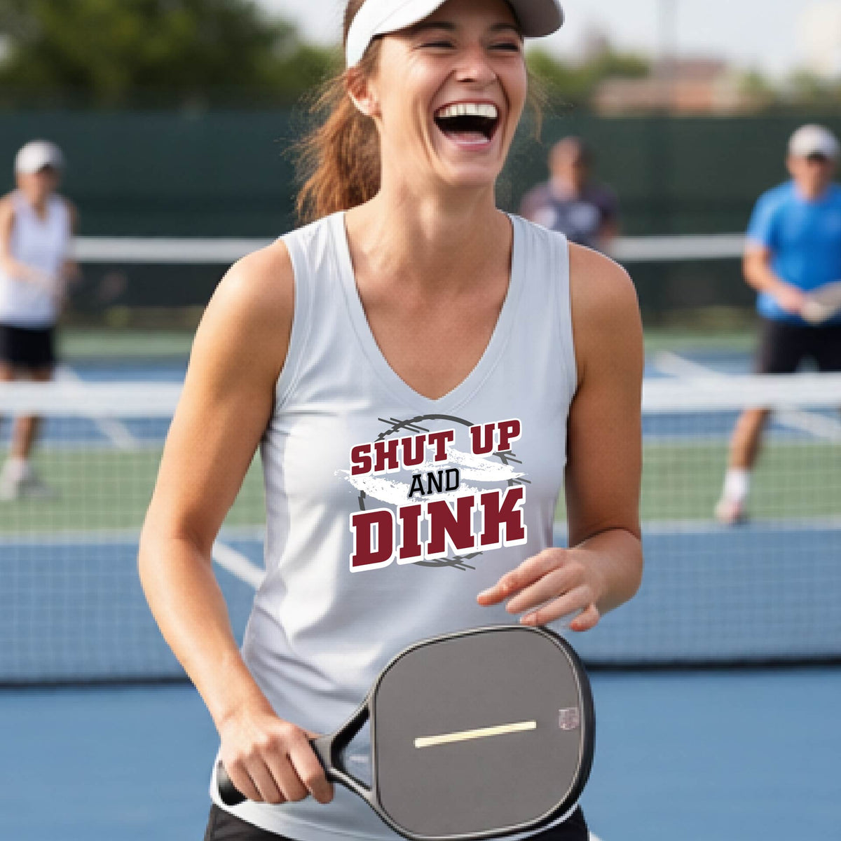 A woman on a pickleball court wearing a silver gray sleeveless t-shirt with a white, gray and black graphic of the text: ‘Shut Up and Dink, standing on a pickleball court.