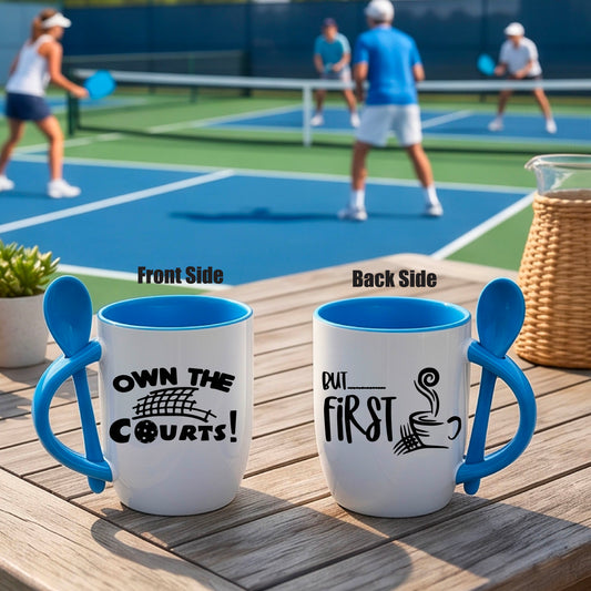 a white and blue 12 ounce coffee mug with a built in spoon in the handle with and a fun coffee cup and pickleball net graphic and wraparound text 'OWN THE COURTS BUT FIRST PICKLEBALL" 
