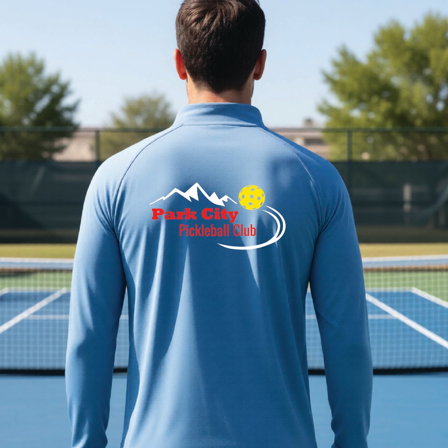 A man viewed from behind wearing a light blue long-sleeve athletic shirt with a 'Park City Pickleball Club' logo printed on the back, standing on an outdoor pickleball court.