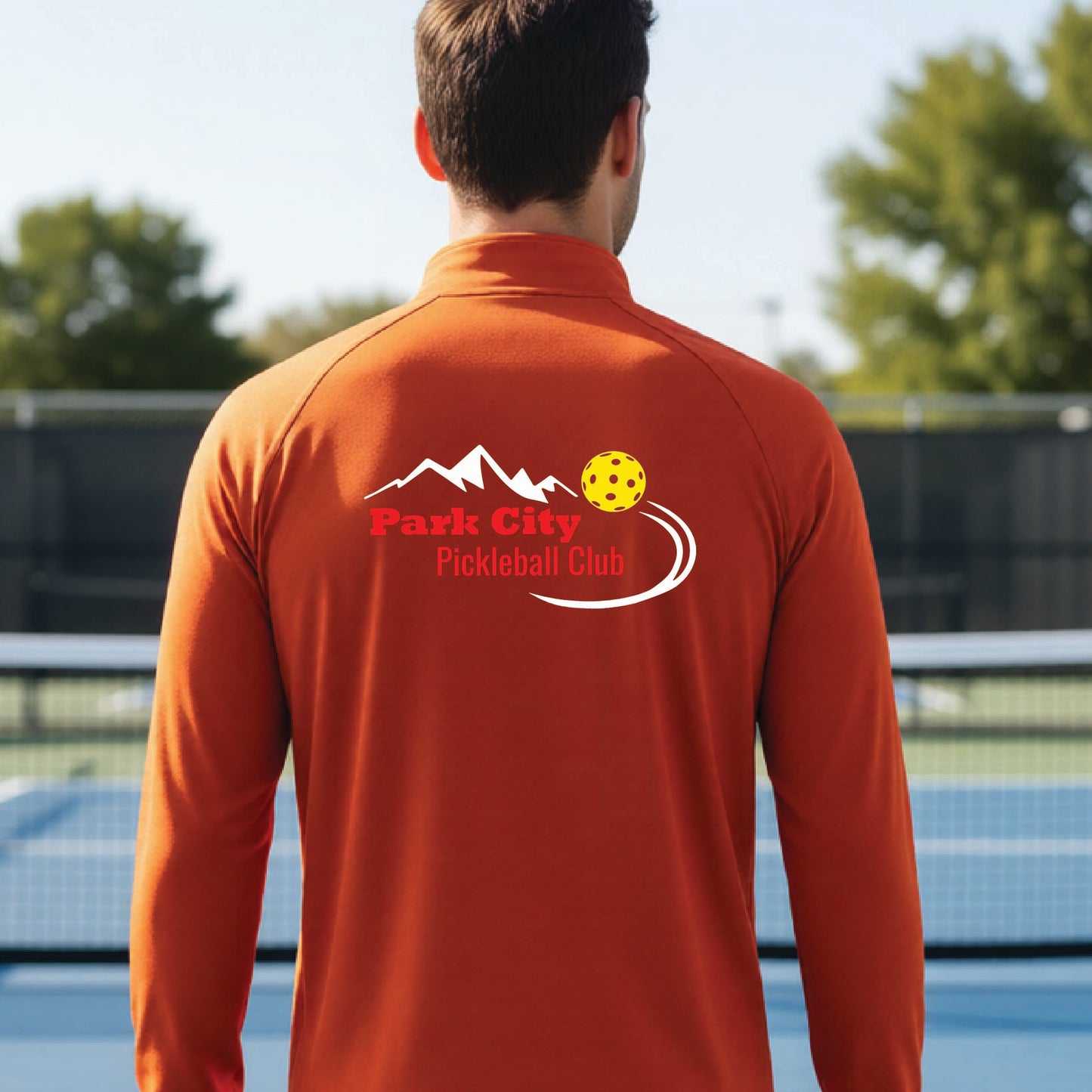 A man viewed from behind wearing a deep orange long-sleeve athletic shirt with a 'Park City Pickleball Club' logo printed on the back, standing on an outdoor pickleball court.