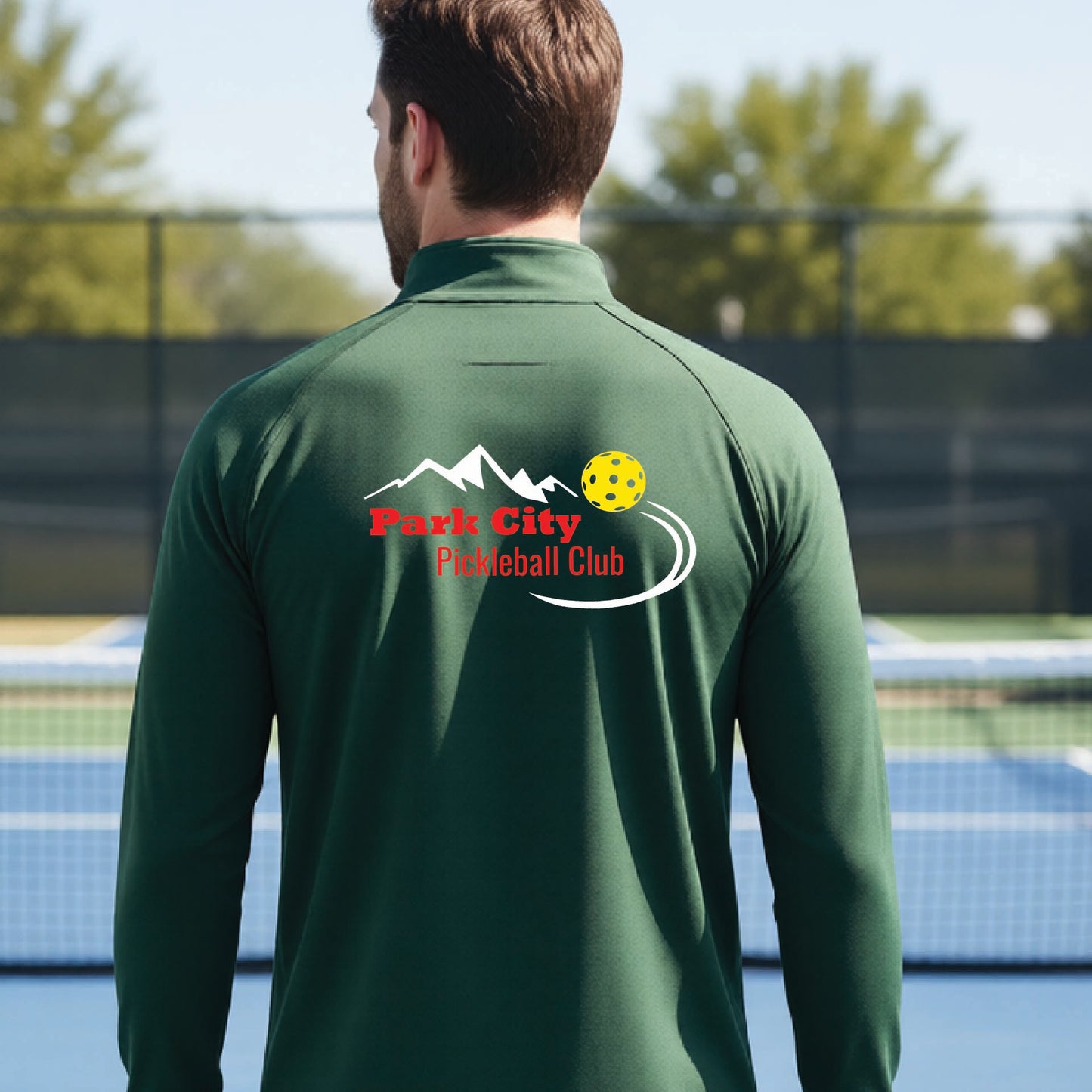 A man viewed from behind wearing a forest green long-sleeve athletic shirt with a 'Park City Pickleball Club' logo printed on the back, standing on an outdoor pickleball court.