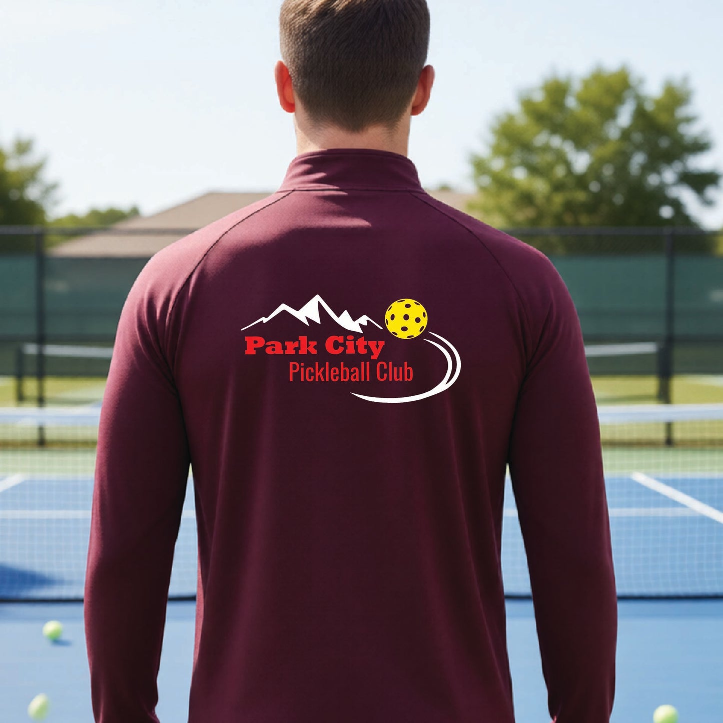 A man viewed from behind wearing a maroon long-sleeve athletic shirt with a 'Park City Pickleball Club' logo printed on the back, standing on an outdoor pickleball court.