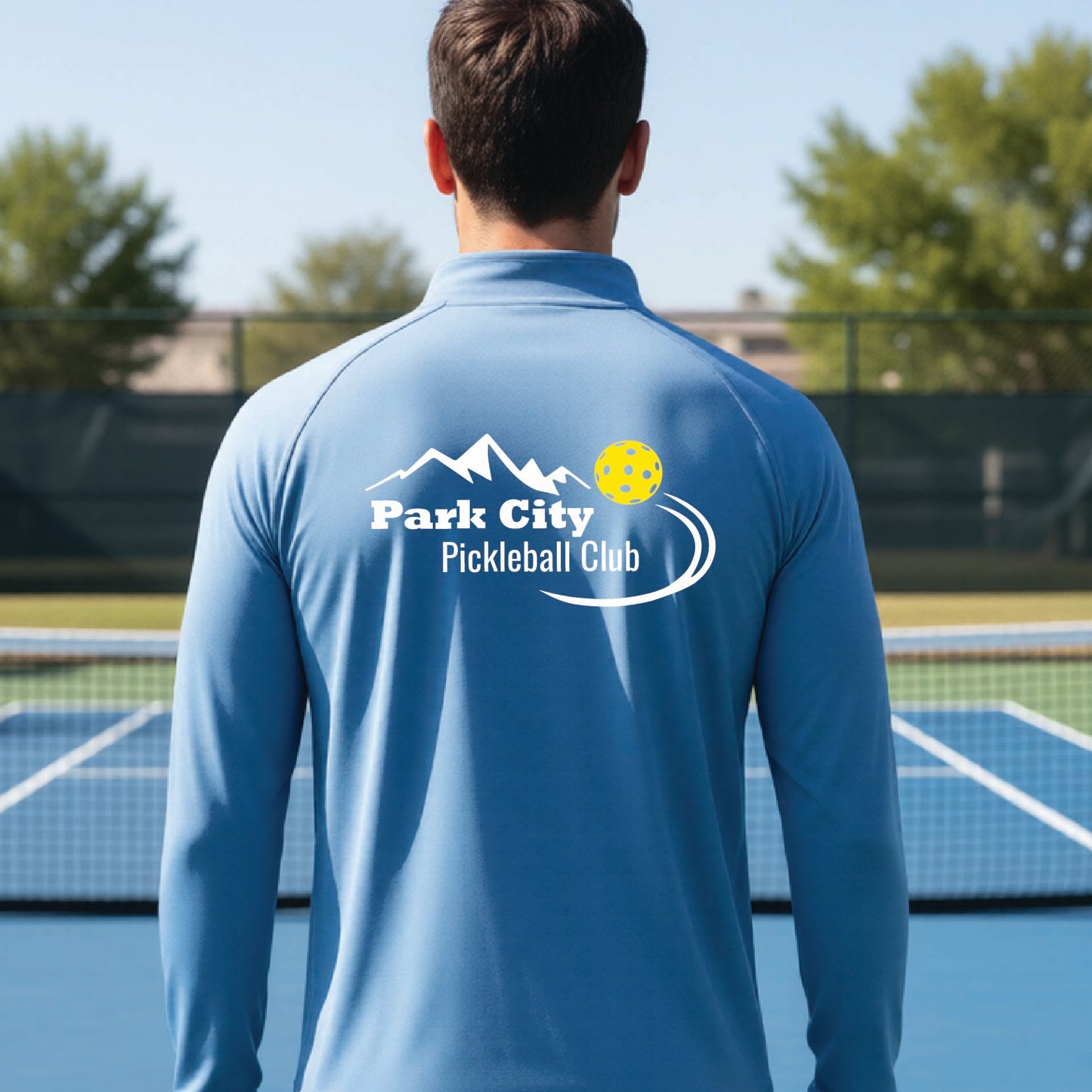 A man viewed from behind wearing a light blue long-sleeve athletic shirt with a 'Park City Pickleball Club' logo printed on the back, standing on an outdoor pickleball court.
