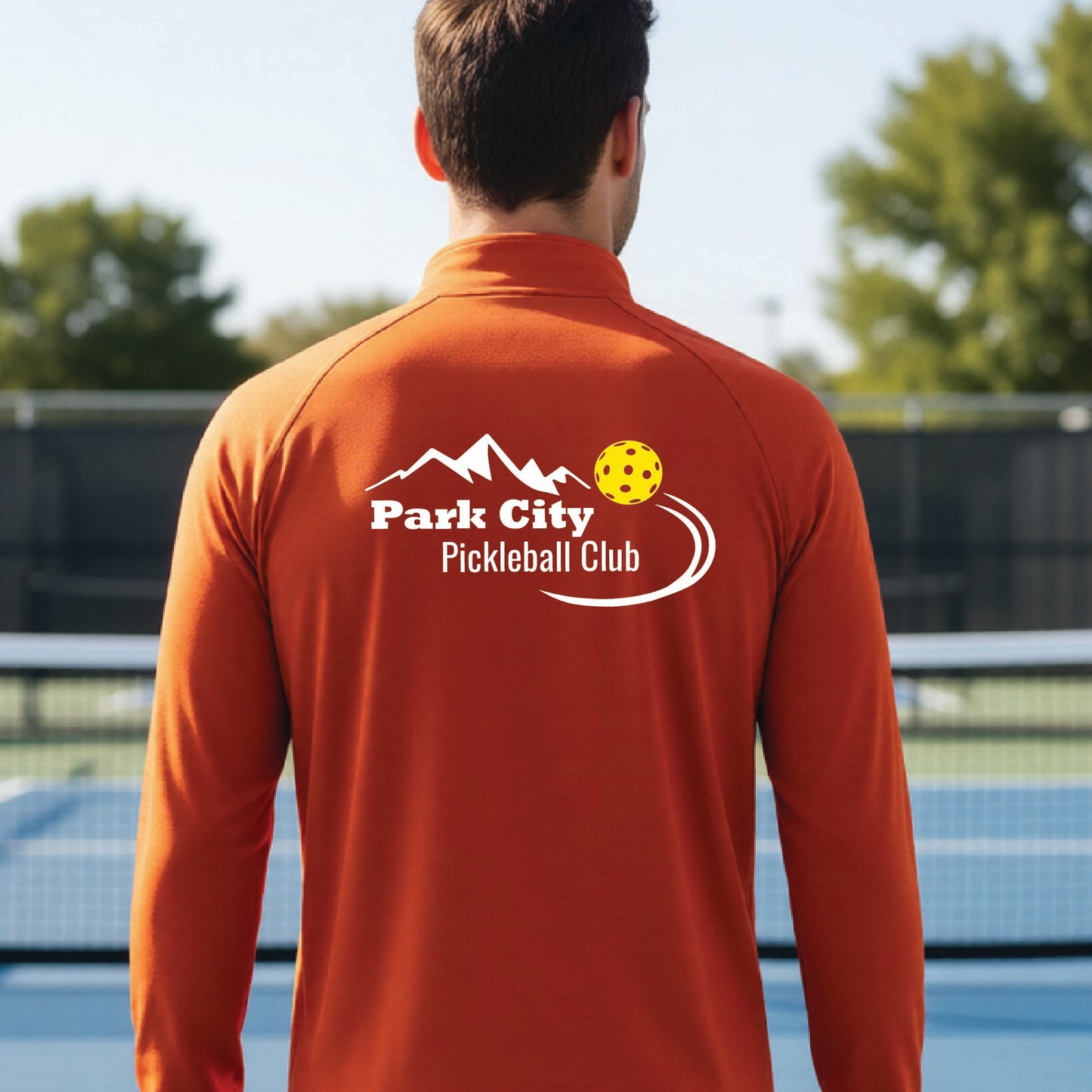 A man viewed from behind wearing a burnt orange long-sleeve athletic shirt with a white 'Park City Pickleball Club' graphic on the back, standing on an outdoor pickleball court.