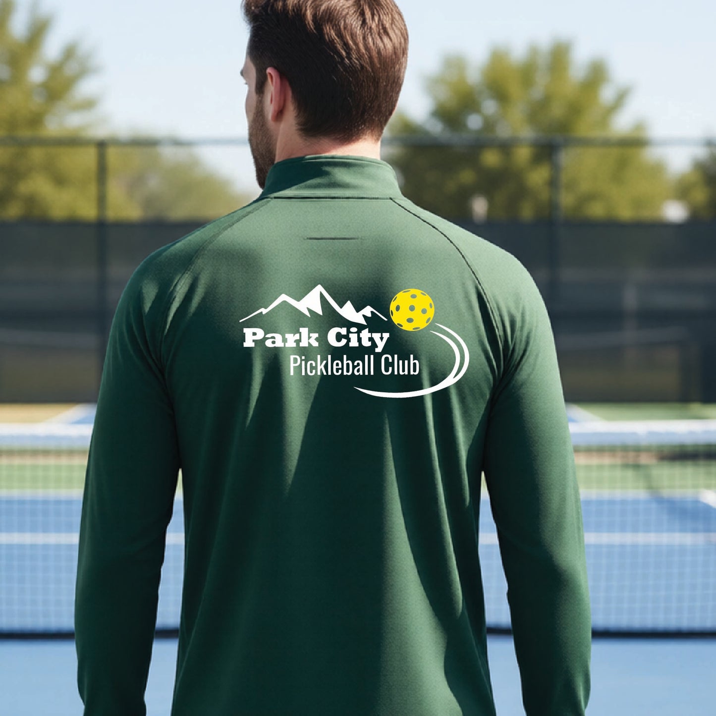 A man viewed from behind wearing a dark green long-sleeve athletic shirt with a 'Park City Pickleball Club' logo printed on the back, standing on an outdoor pickleball court.