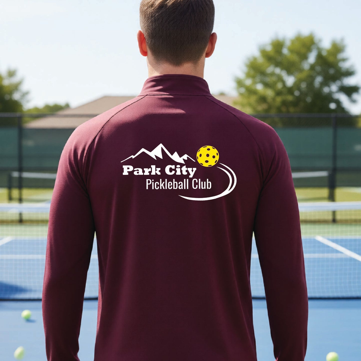 A man viewed from behind wearing a maroon long-sleeve athletic quarter-zip pullover with a white 'Park City Pickleball Club' graphic printed on the back, standing on a blue pickleball court.