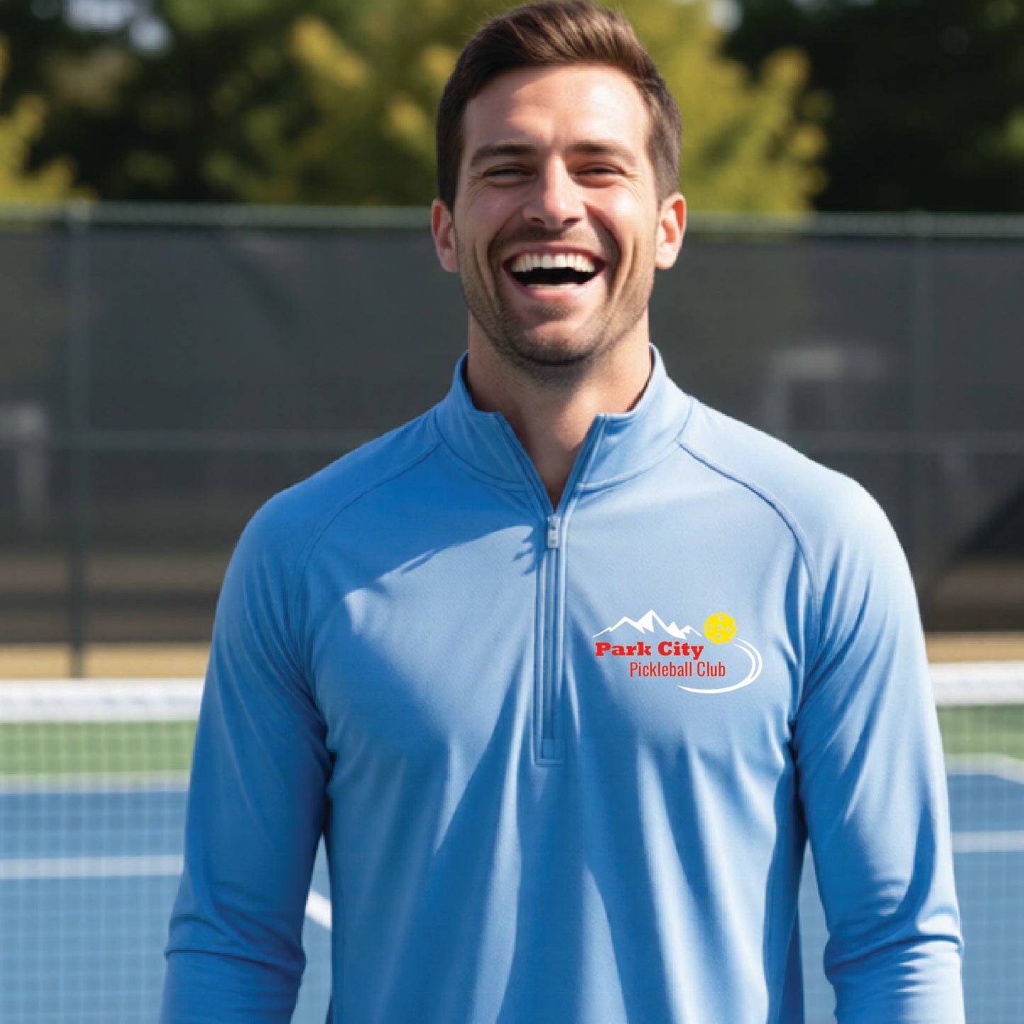 A smiling man wearing a light blue long-sleeve athletic quarter-zip pullover with a 'Park City Pickleball Club' logo on the left chest, standing on an outdoor pickleball court.