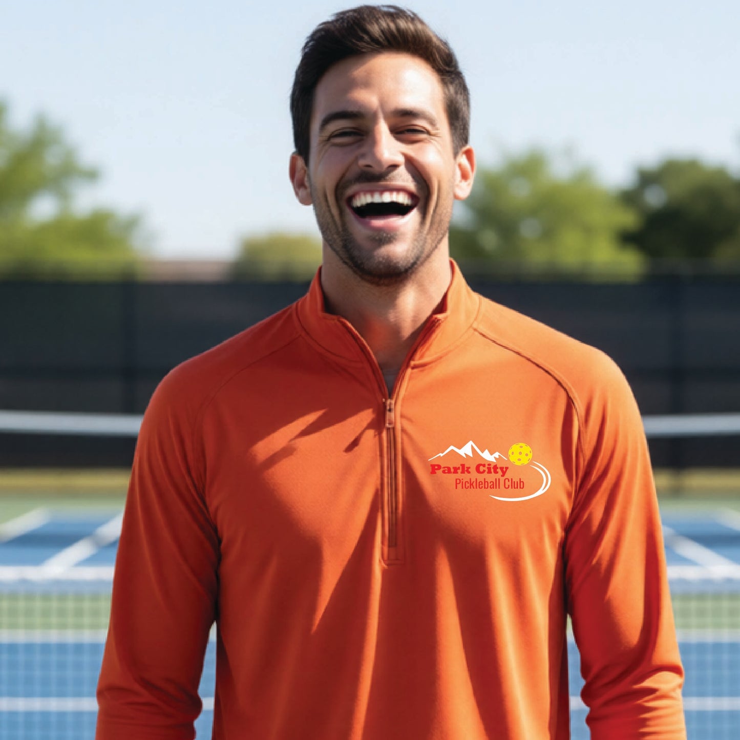 A smiling man wearing a burnt orange long-sleeve athletic quarter-zip pullover with a 'Park City Pickleball Club' logo on the left chest, standing on an outdoor pickleball court.