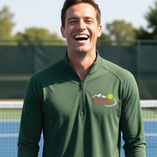 A smiling man wearing a forest green long-sleeve athletic quarter-zip pullover with a 'Park City Pickleball Club' logo on the left chest, standing on an outdoor pickleball court.