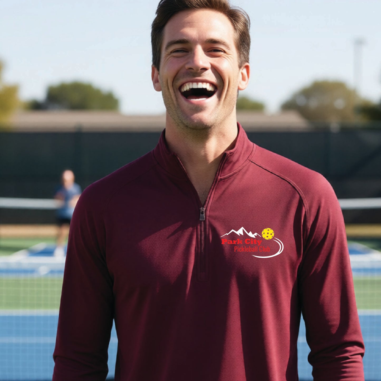A smiling man wearing a maroon long-sleeve athletic quarter-zip pullover with a 'Park City Pickleball Club' logo on the left chest, standing on an outdoor pickleball court.