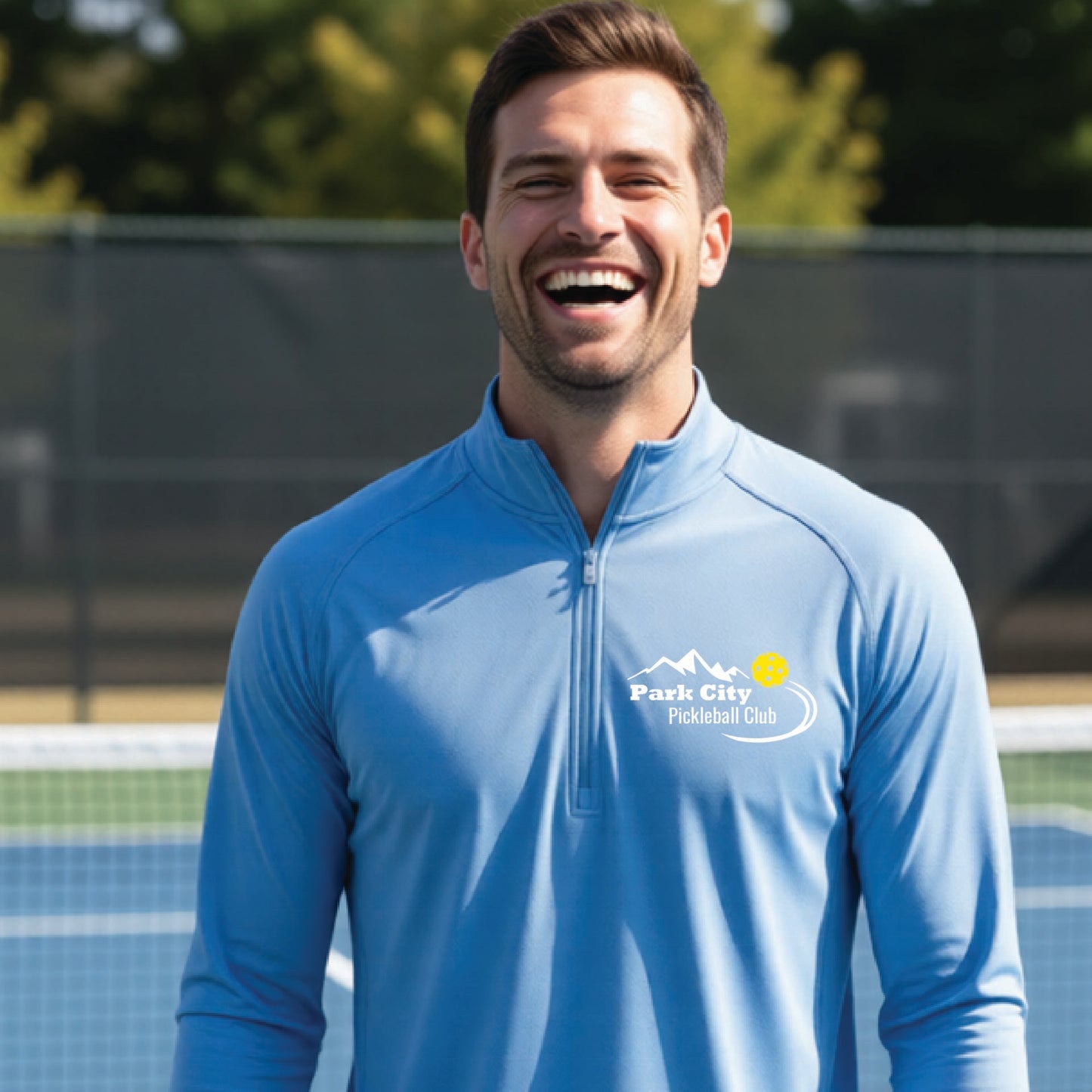 A smiling man wearing a light blue long-sleeve athletic quarter-zip pullover with a 'Park City Pickleball Club' logo on the left chest, standing on an outdoor pickleball court.
