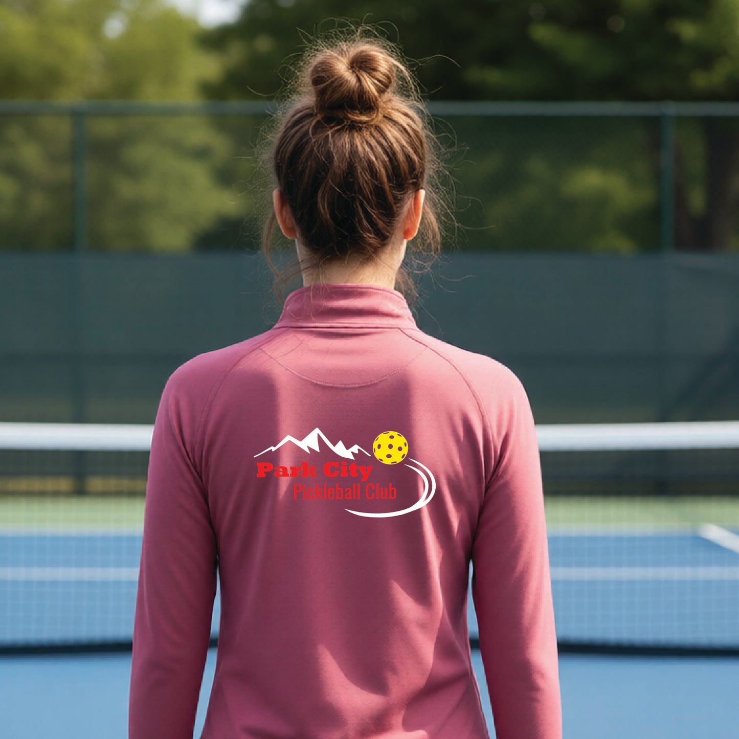 A woman viewed from behind on an outdoor pickleball court, wearing a dusty rose long-sleeve quarter-zip athletic top with a custom 'Park City Pickleball Club' logo printed on the back.