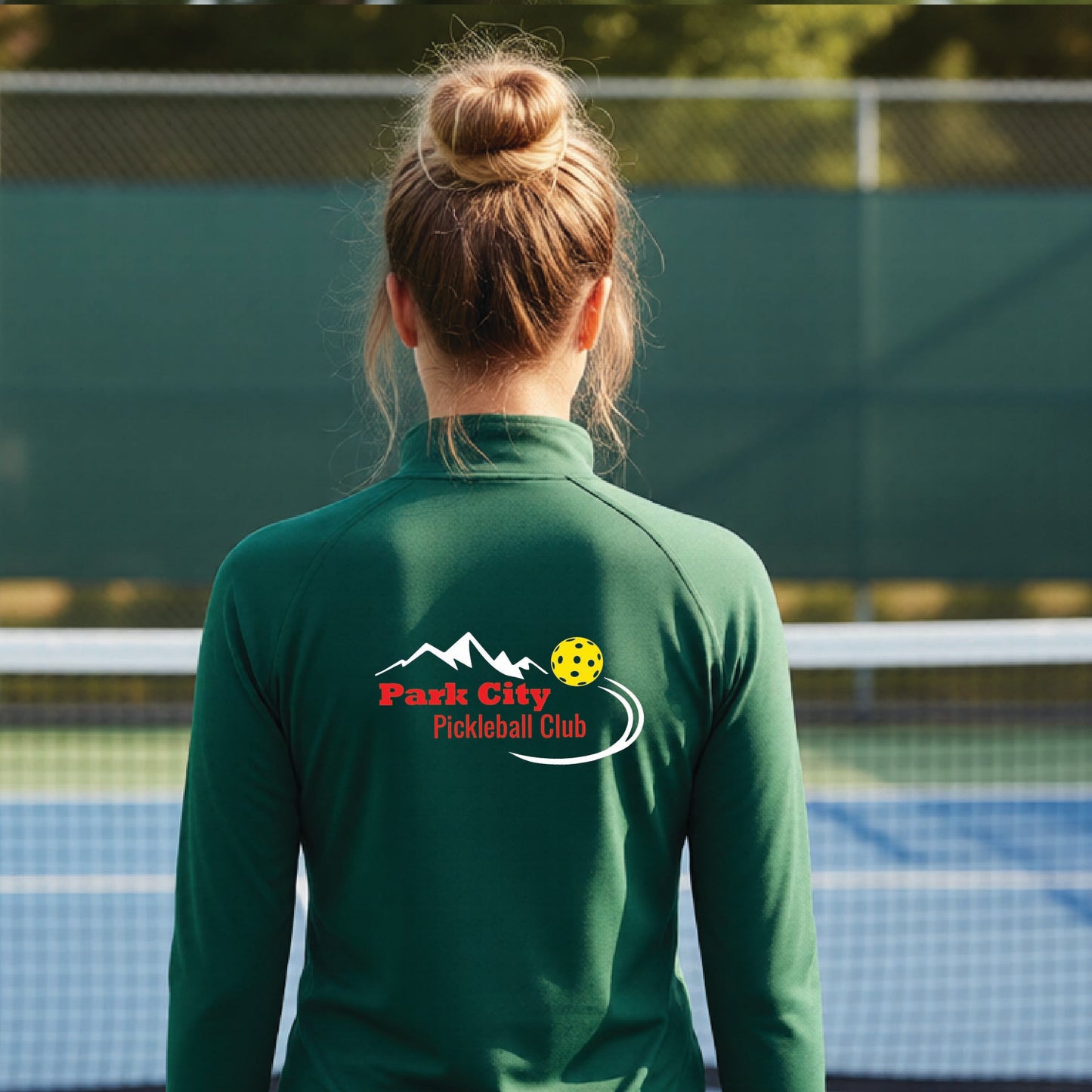 A woman viewed from behind on an outdoor pickleball court, wearing a forest green long-sleeve quarter-zip athletic top with a custom 'Park City Pickleball Club' logo printed on the back.