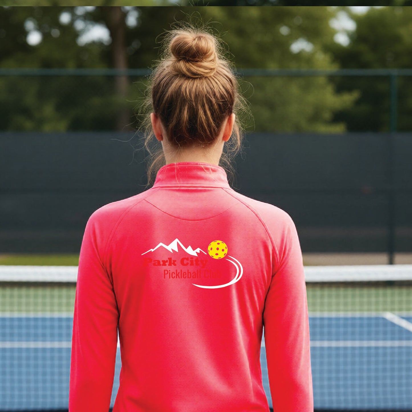 A woman viewed from behind on an outdoor pickleball court, wearing a hot coral pink long-sleeve quarter-zip athletic top with a custom 'Park City Pickleball Club' logo printed on the back.