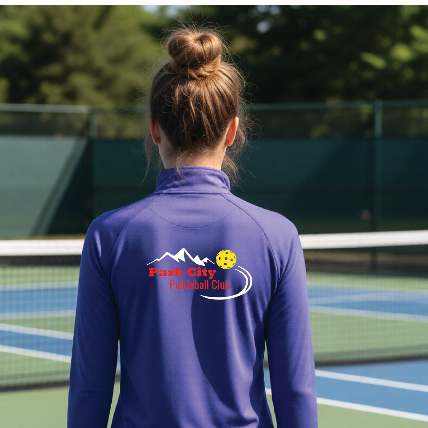 A woman viewed from behind on an outdoor pickleball court, wearing a purple long-sleeve quarter-zip athletic top with a custom 'Park City Pickleball Club' logo printed on the back.