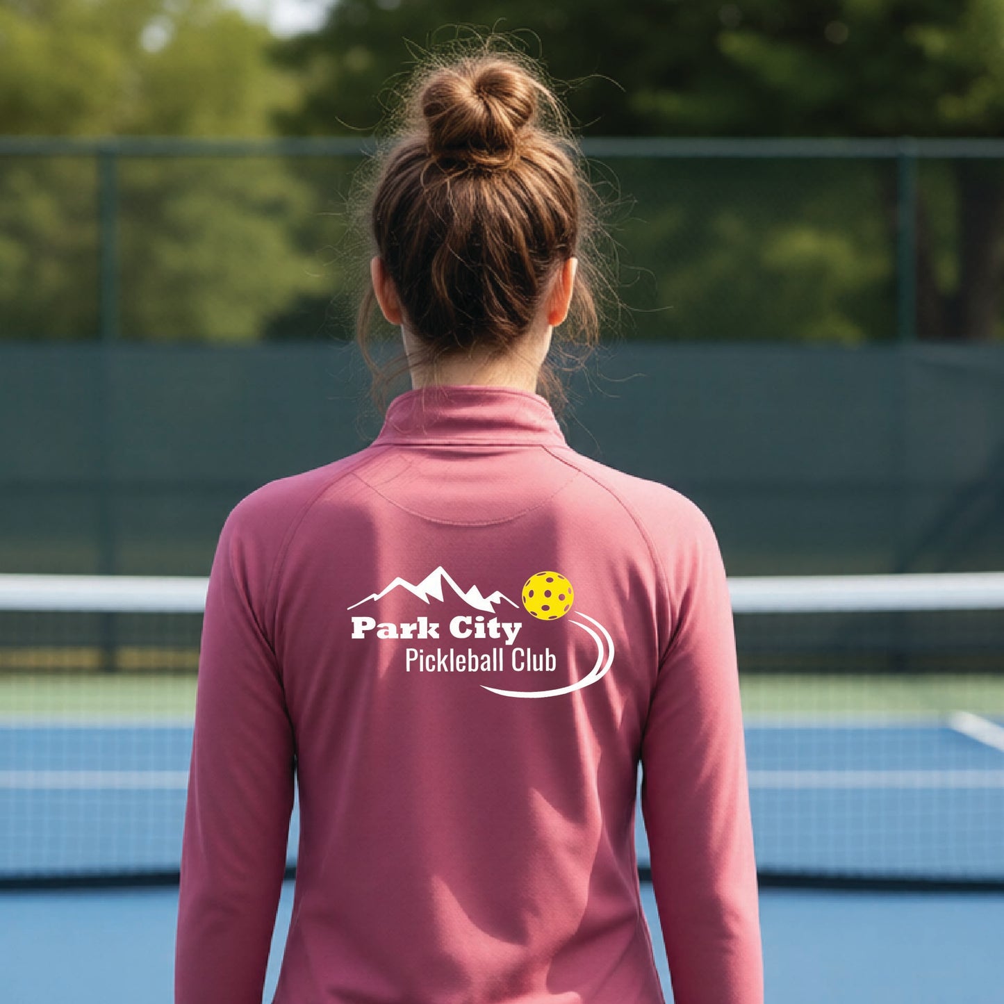 A woman viewed from behind on an outdoor pickleball court, wearing a dusty rose long-sleeve quarter-zip athletic top with a custom 'Park City Pickleball Club' logo printed on the back.