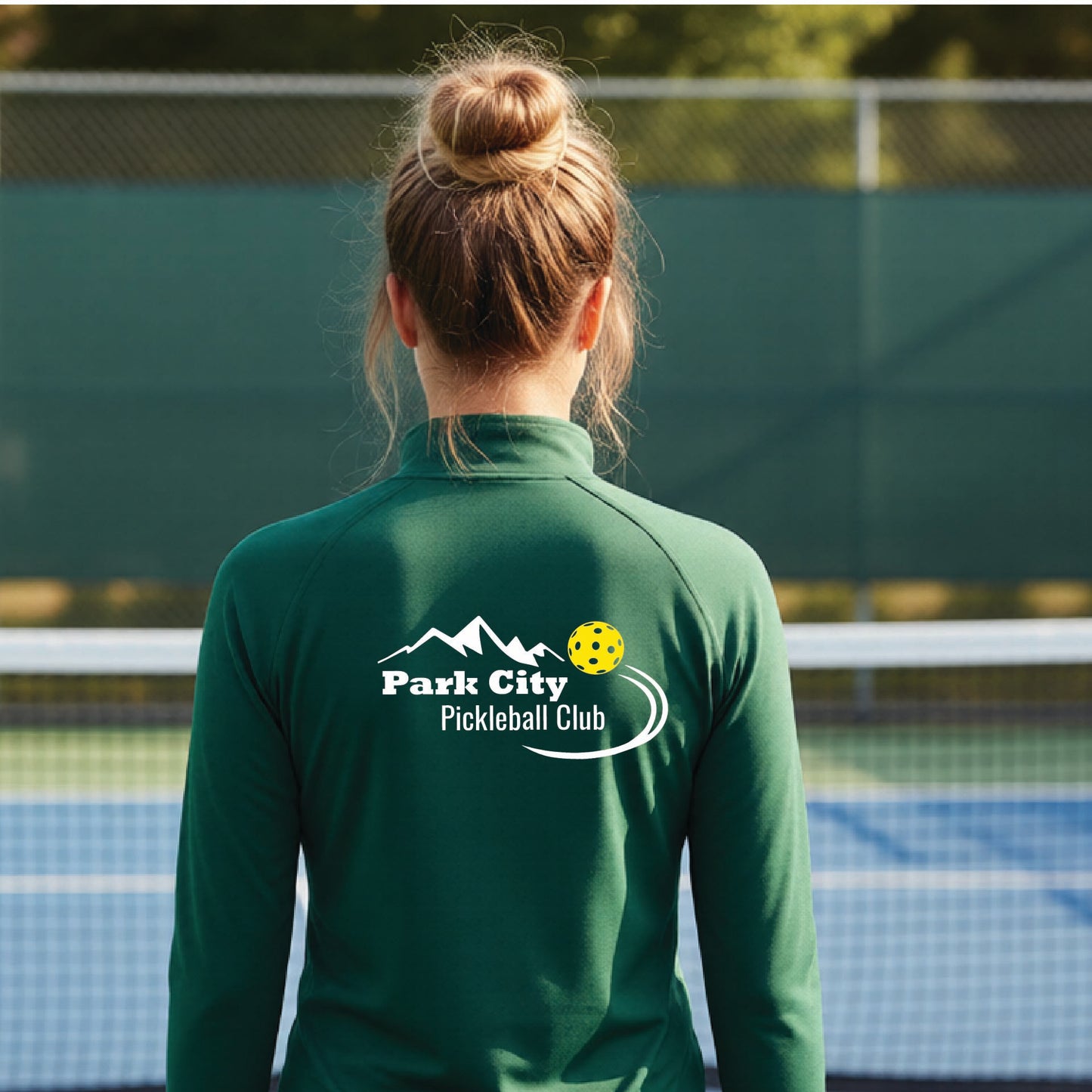 A woman viewed from behind on an outdoor pickleball court, wearing a forest green long-sleeve quarter-zip athletic top with a custom 'Park City Pickleball Club' logo printed on the back.