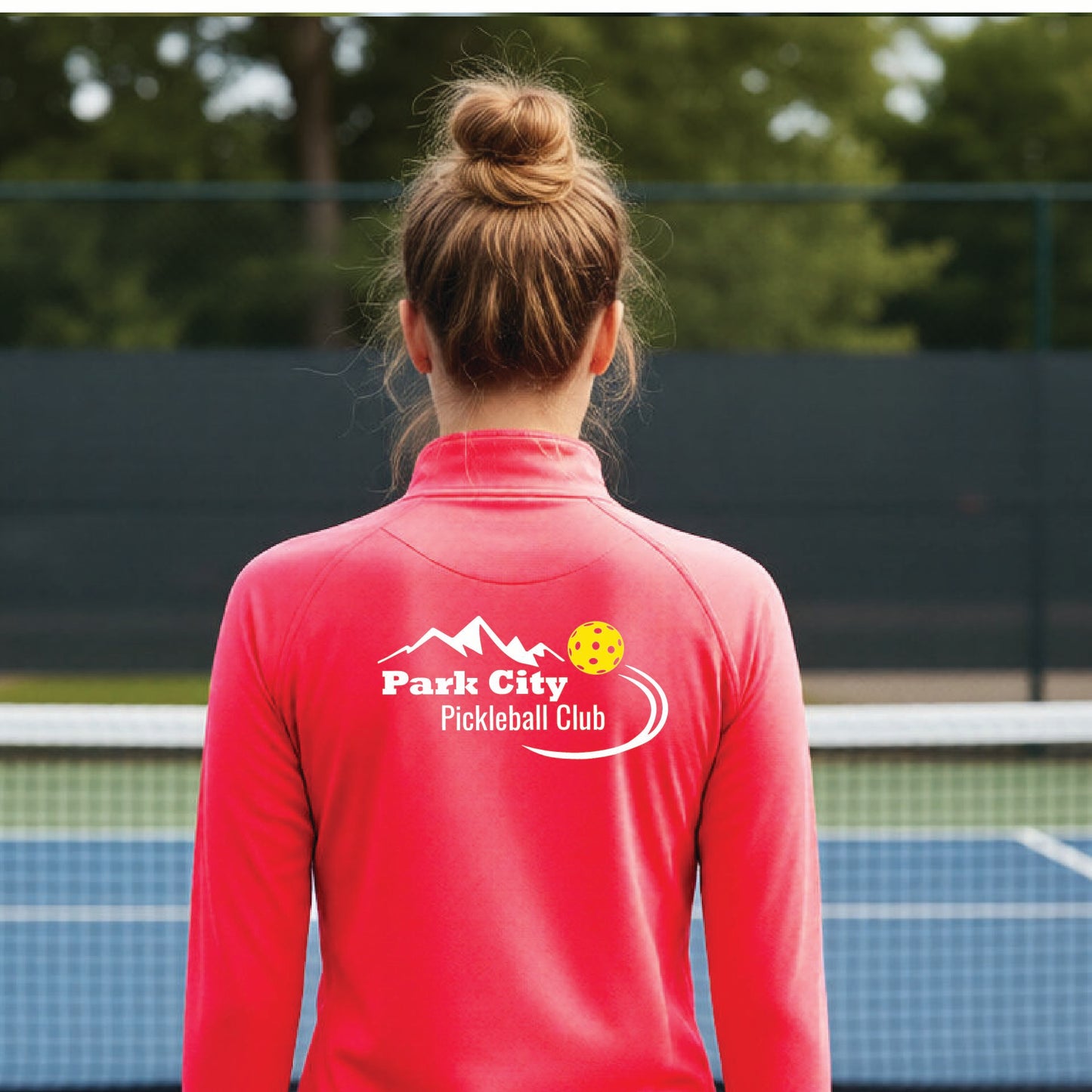 A woman viewed from behind on an outdoor pickleball court, wearing a  hot coral pink long-sleeve quarter-zip athletic top with a custom 'Park City Pickleball Club' logo printed on the back.