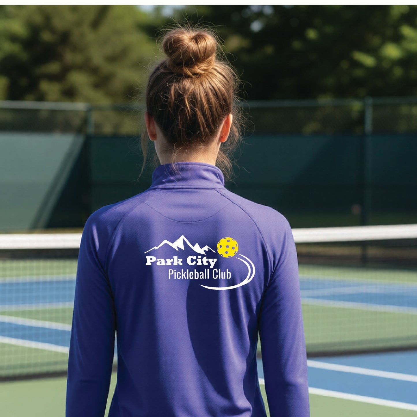 A woman viewed from behind on an outdoor pickleball court, wearing a purple long-sleeve quarter-zip athletic top with a custom 'Park City Pickleball Club' logo printed on the back.