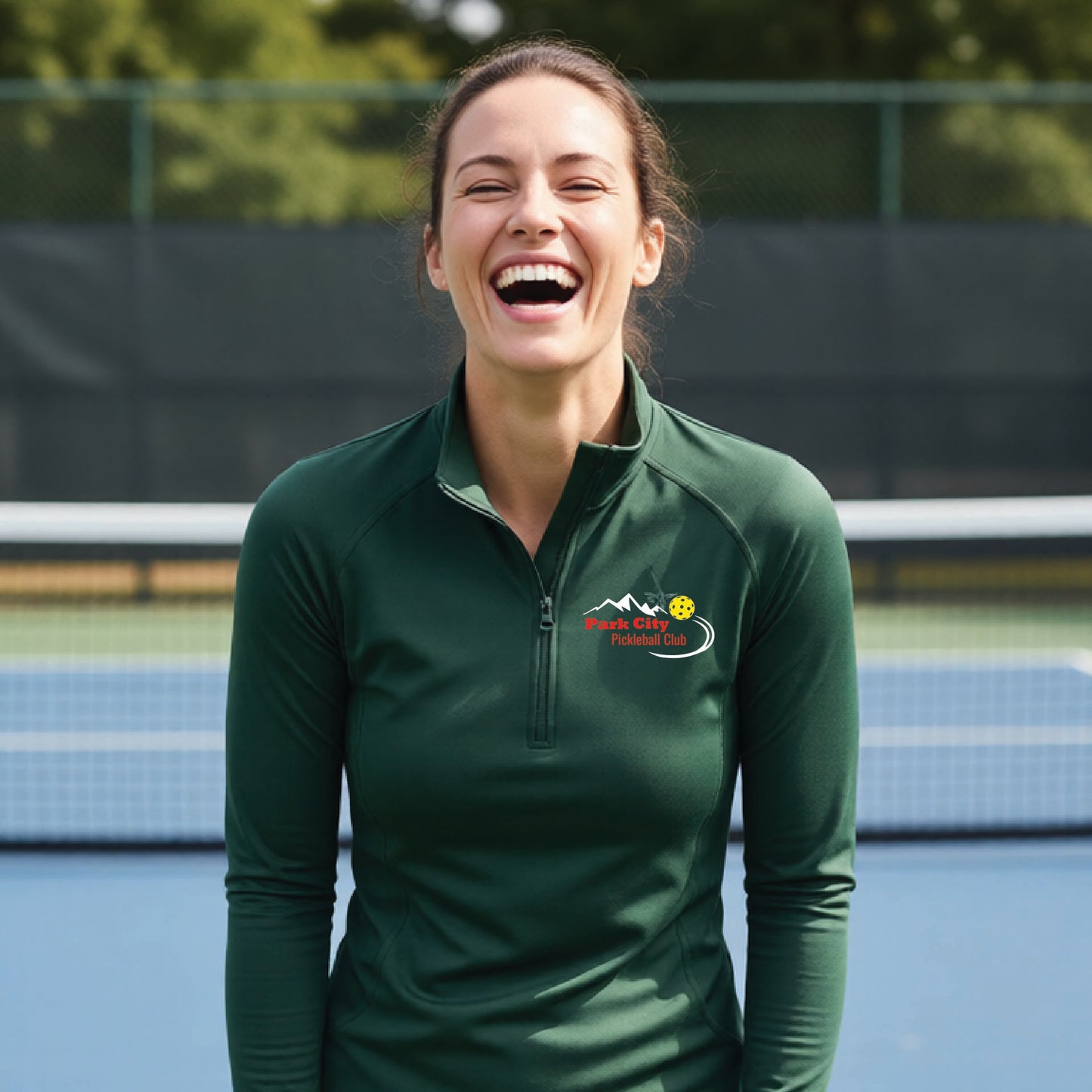 A woman on an outdoor pickleball court, wearing a forest green long-sleeve quarter-zip athletic top with a custom 'Park City Pickleball Club' logo printed on the pocket area.