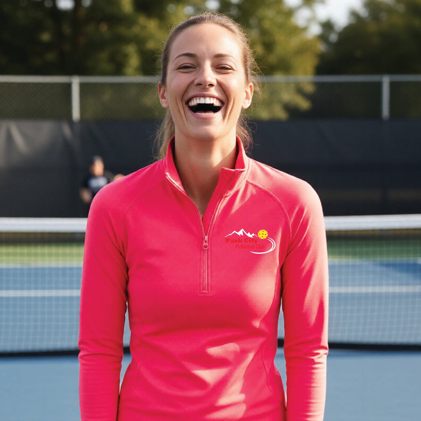 A woman on an outdoor pickleball court, wearing a hot coral pink long-sleeve quarter-zip athletic top with a custom 'Park City Pickleball Club' logo printed on the pocket area.